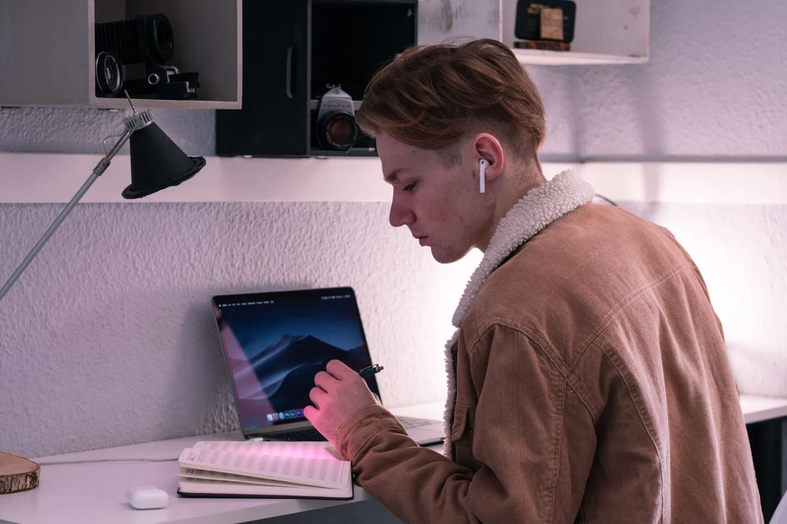 Side view of serious young male student in TWS earbuds writing in planner while sitting at desk with laptop